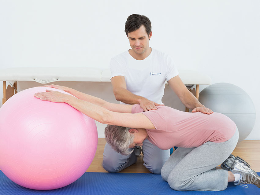 Eine ältere Dame arbeitet bei der Physiotherapie mit einem rosa Gymnastikball